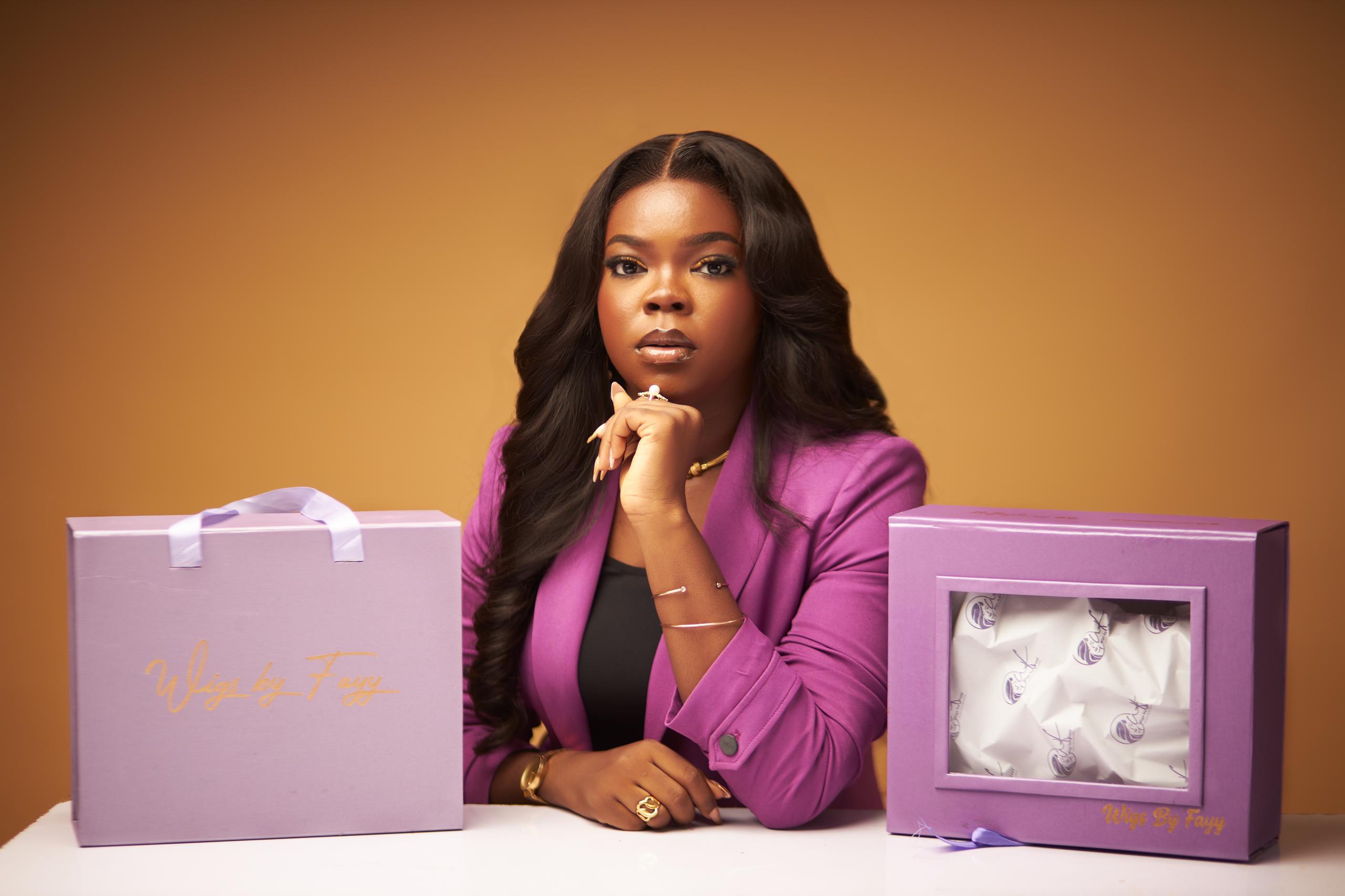 Model wearing Wigs by Fayy seated between two signature purple gift boxes